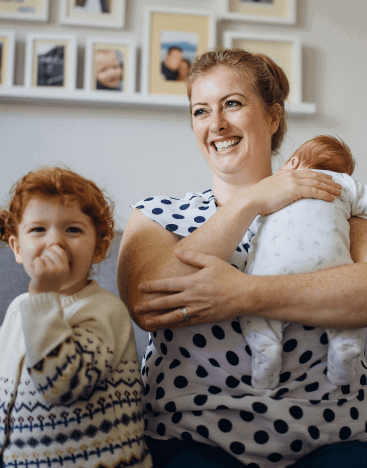 A mother and her 2 small children, she is holding the smallest baby in her arms whilst smiling, her elder child is smiling at the camera